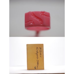 May include: A pink wax ring with a detailed design. The ring is sitting on a brown box with the text "In Bins Kock Chappell CHALLAN" written on it.