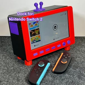 May include: A red and black Nintendo Switch 2 dock, designed as a retro TV. The screen shows a game menu. Two black controllers with blue and orange details are in front. The text "Dock for: Nintendo Switch 2" is displayed on the dock.