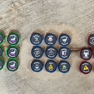 May include: A set of 18 round tokens with various symbols and text. The tokens are black with white text and symbols. The tokens are arranged in three rows of six. The tokens are on a wooden surface.