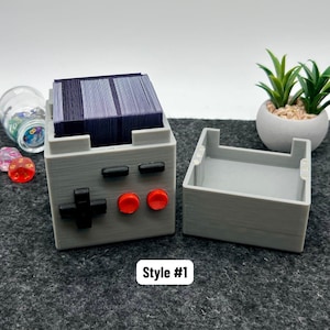 May include: A gray 3D-printed deck box shaped like a handheld gaming console, filled with a stack of dark blue cards. The box features a black directional pad, two red circular buttons, and two black rectangular buttons. Text reads Style #1.