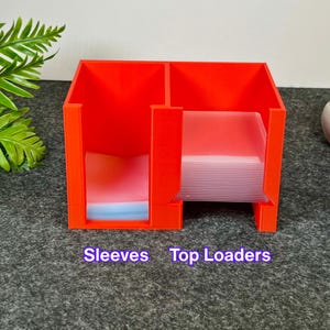 May include: A bright red plastic storage organiser with two distinct vertical compartments. The left section is labelled Sleeves and contains clear protective sleeves, while the right section is labelled Top Loaders and holds clear rigid top loaders.