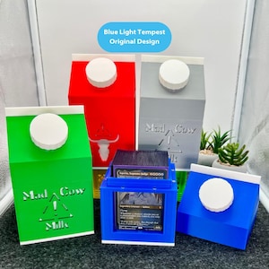May include: A collection of colorful card storage boxes shaped like milk cartons. The boxes are in green, red, gray, and blue, each with a white cap. The text "Mad Cow Milk" is visible on the green box. The text "Blue Light Tempest Original Design" is also visible.