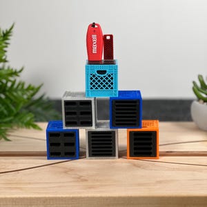 May include: A display of colorful, geometric storage cubes in blue, orange, gray, and white, arranged on a wooden surface. A small blue crate supports a red Maxell USB drive and a dark red USB drive. A fern and a small plant are in the background.