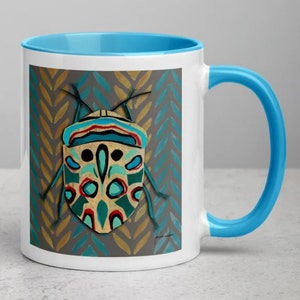 May include: White ceramic mug with a light blue interior and handle. The mug features a colorful illustration of a stylized insect with teal, red, and gold accents against a gray background with a chevron pattern. The mug has a glossy finish.