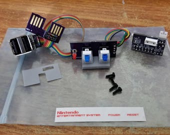 May include: A set of electronic components for a game console including a black circuit board with two blue switches, a USB port module with rainbow ribbon cables, a small black connector board, three black screws, a grey plastic piece, and a label.