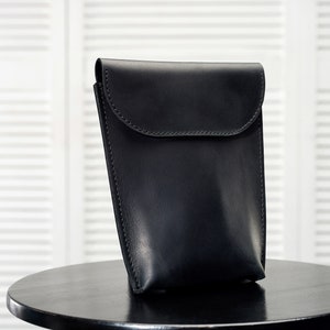 Puede incluir: Una funda de tel&eacute;fono de cuero negro con cierre de solapa. La funda est&aacute; sobre una mesa negra.