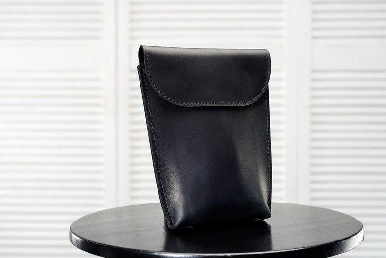 Puede incluir: Una funda de tel&eacute;fono de cuero negro con cierre de solapa. La funda est&aacute; sobre una mesa negra.