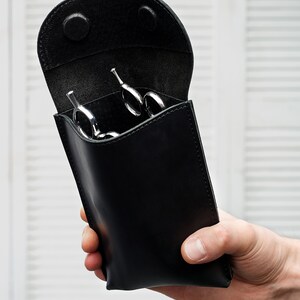 Puede incluir: Estuche de cuero negro con una solapa de apertura, que contiene varias tijeras de peluquer&iacute;a plateadas.