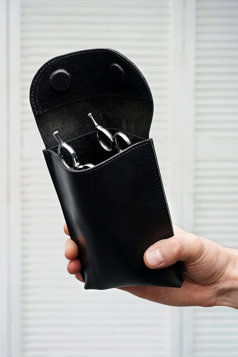 Puede incluir: Estuche de cuero negro con una solapa de apertura, que contiene varias tijeras de peluquer&iacute;a plateadas.