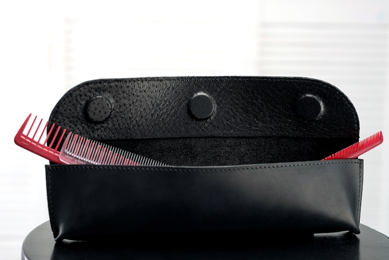 Puede incluir: Una funda de cuero negro con tres broches, abierta para revelar dos peines rojos y transparentes en el interior.