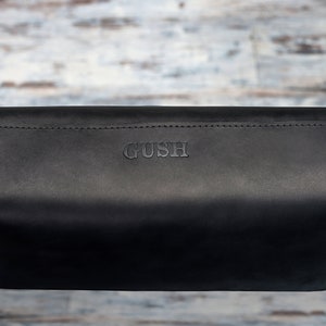 Puede incluir: Bolsa de cuero negro con la palabra "GUSH" grabada en la parte delantera.