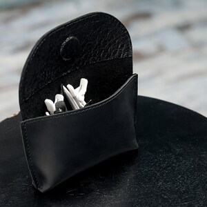 Puede incluir: Una funda de cuero negro con cierre de presi&oacute;n, abierta para revelar un par de auriculares blancos en su interior.