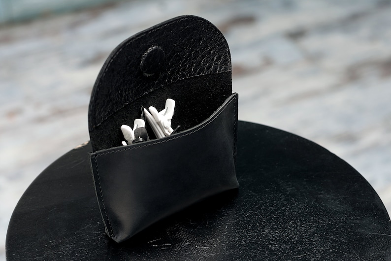 Puede incluir: Una funda de cuero negro con cierre de presi&oacute;n, abierta para revelar un par de auriculares blancos en su interior.