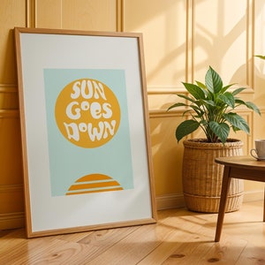 Peut inclure: Une impression encadrée avec un fond bleu clair et un cercle jaune avec le texte "SUN GOES DOWN" en blanc. L'impression est accrochée à un mur jaune avec des lambris en bois.