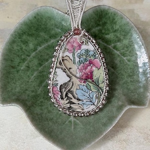 May include: A silver wire wrapped pendant featuring a vintage floral pattern on a white background. The pendant is oval shaped and has a silver bead border.