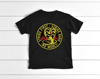 May include: Black t-shirt with a yellow circle graphic featuring a cobra with the text "Strike First Strike Hard No Mercy" and "Cobra Kai" in red.