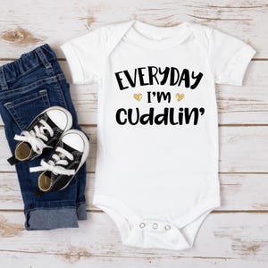 May include: White baby onesie with black text that reads "EVERYDAY I'M CUDDLIN'" with two gold hearts above the word "I'M". The onesie is paired with blue jeans and black sneakers.