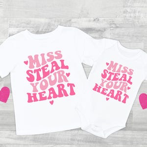 Pode incluir: Duas camisas brancas com texto rosa que diz "Miss Steal Your Heart" com um coração rosa de cada lado do texto. As camisas estão sobre um fundo de madeira clara.