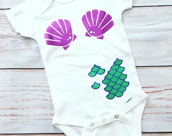 Peut inclure: Combinaison blanche pour bébé avec un motif de coquillage violet sur le devant et une queue de sirène verte sur le bas. La combinaison porte le logo de la marque Gerber sur le devant.