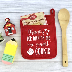 May include: Red oven mitt with a pocket that says "Thanks for making me one smart cookie" with a chocolate chip cookie mix packet inside. There is a wooden schoolhouse, a yellow pencil, and a wooden spoon in the background.