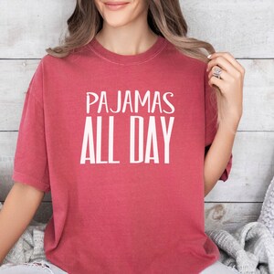 a woman sitting on a bed wearing a red shirt that says pajamas all day