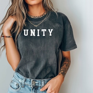 May include: A dark gray t-shirt with the word "UNITY" printed in white on the front. The shirt is worn by a person with long brown hair and a gold chain necklace.