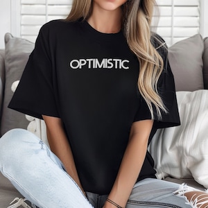 May include: A black oversized t-shirt with the word "OPTIMISTIC" printed in white on the front. The shirt is being worn by a person with long blonde hair.