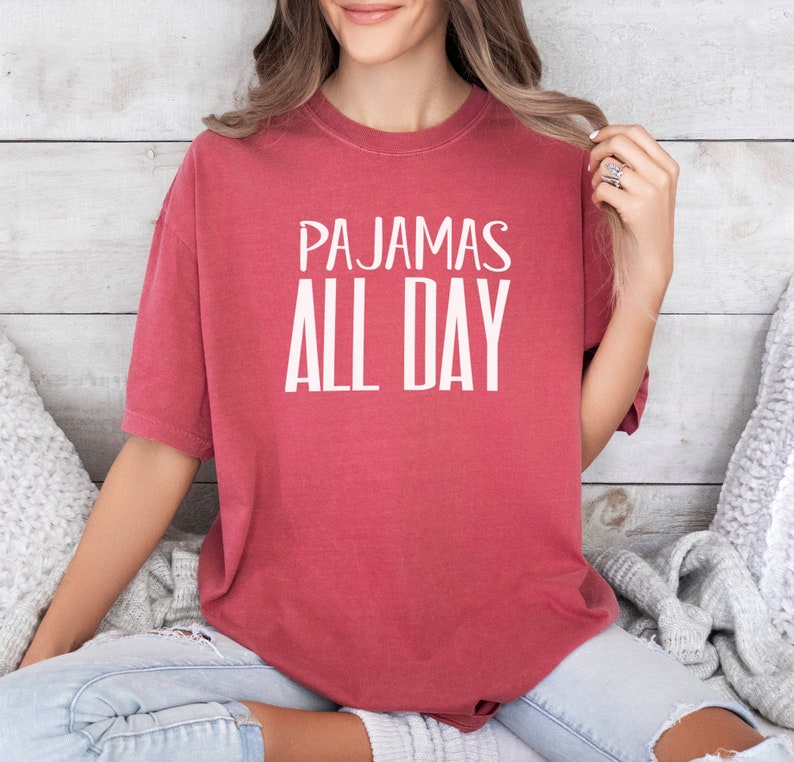 a woman sitting on a bed wearing a red shirt that says pajamas all day