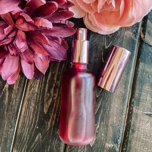 K&ouml;nnte beinhalten: Eine rote Glasflasche mit einem ros&eacute;goldenen Spr&uuml;hkopf und Verschluss. Die Flasche steht auf einer Holzoberfl&auml;che mit rosa und burgunderfarbenen Blumen.