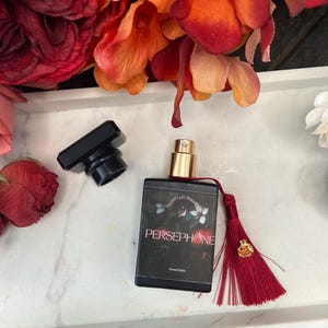 K&ouml;nnte beinhalten: Eine schwarze Glasflasche mit Parf&uuml;m, einem goldenen Verschluss und einem roten Quast. Die Flasche ist mit "Secret Lore Botanicals" und "Persephone" sowie einem floralen Design beschriftet.