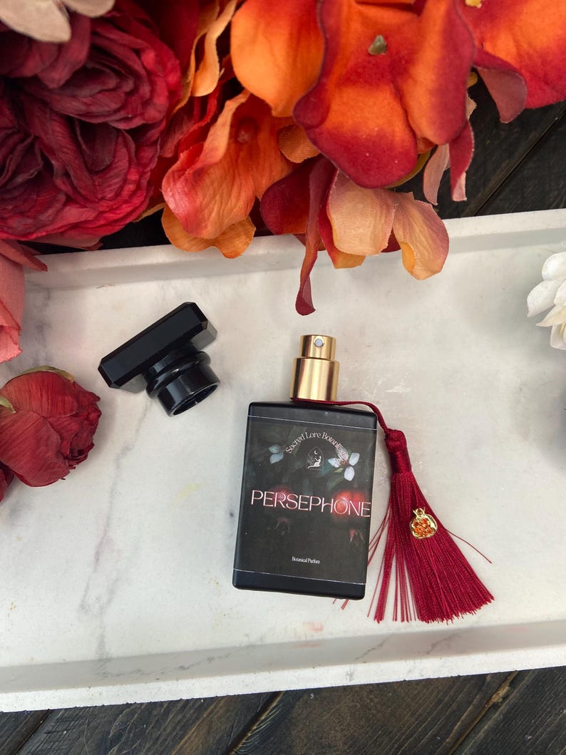 K&ouml;nnte beinhalten: Eine schwarze Glasflasche mit Parf&uuml;m, einem goldenen Verschluss und einem roten Quast. Die Flasche ist mit "Secret Lore Botanicals" und "Persephone" sowie einem floralen Design beschriftet.
