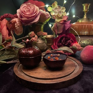 Puede incluir: Dos peque&ntilde;os recipientes en forma de granada de color rojo oscuro con dise&ntilde;os intrincados. Un recipiente est&aacute; lleno de cera naranja y roja. Los recipientes est&aacute;n sobre una base de madera con un acabado marr&oacute;n oscuro. Los recipientes est&aacute;n rodeados de rosas rojas, un melocot&oacute;n y un libro.