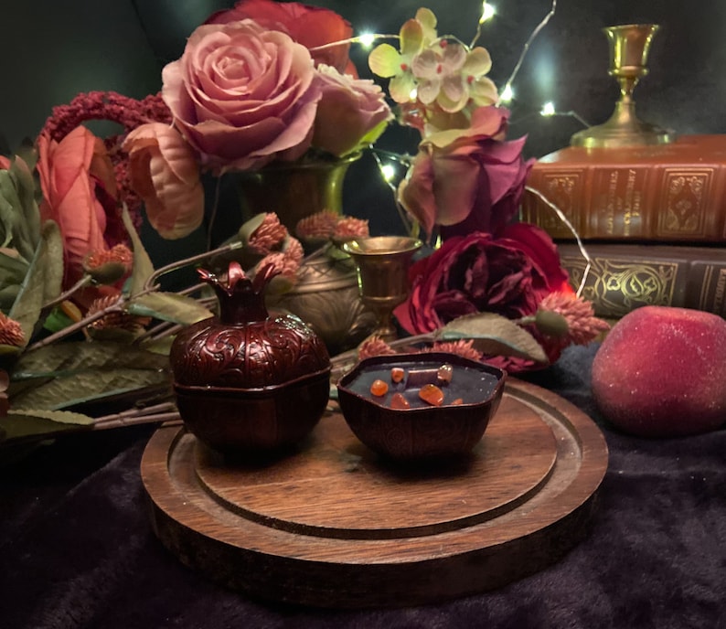 Puede incluir: Dos peque&ntilde;os recipientes en forma de granada de color rojo oscuro con dise&ntilde;os intrincados. Un recipiente est&aacute; lleno de cera naranja y roja. Los recipientes est&aacute;n sobre una base de madera con un acabado marr&oacute;n oscuro. Los recipientes est&aacute;n rodeados de rosas rojas, un melocot&oacute;n y un libro.