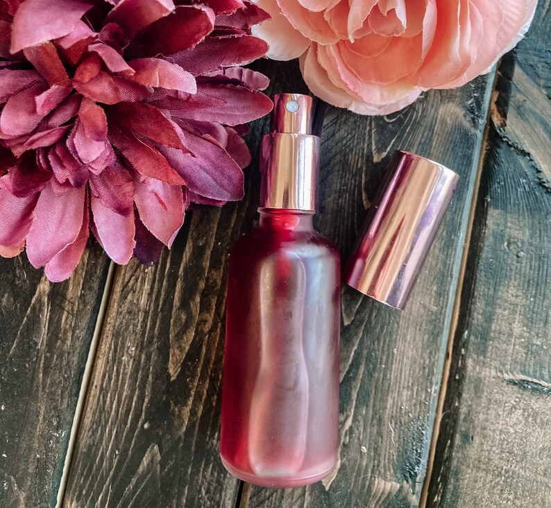 K&ouml;nnte beinhalten: Eine rote Glasflasche mit einem ros&eacute;goldenen Spr&uuml;hkopf und Verschluss. Die Flasche steht auf einer Holzoberfl&auml;che mit rosa und burgunderfarbenen Blumen.