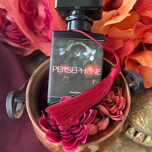 K&ouml;nnte beinhalten: Eine kleine schwarze Parf&uuml;mflasche mit einem roten Quast. Die Flasche ist mit "Sacred Lore Botanicals" und "PERSEPHONE" beschriftet. Die Flasche steht in einer Kupferschale mit roten Blumen.