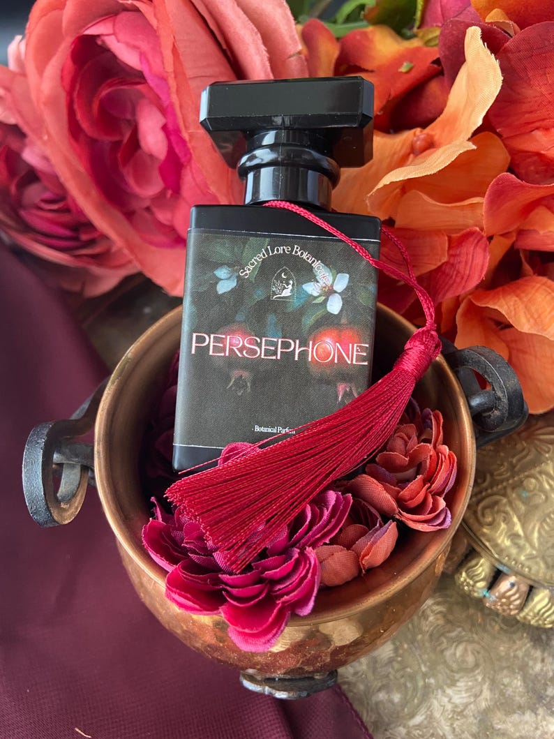 K&ouml;nnte beinhalten: Eine kleine schwarze Parf&uuml;mflasche mit einem roten Quast. Die Flasche ist mit "Sacred Lore Botanicals" und "PERSEPHONE" beschriftet. Die Flasche steht in einer Kupferschale mit roten Blumen.