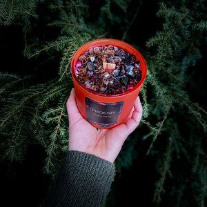 May include: A red candle jar with a black and brown wax blend and a wooden wick. The jar has a label that says "PHOENIX" with the words "with every single soul" underneath.