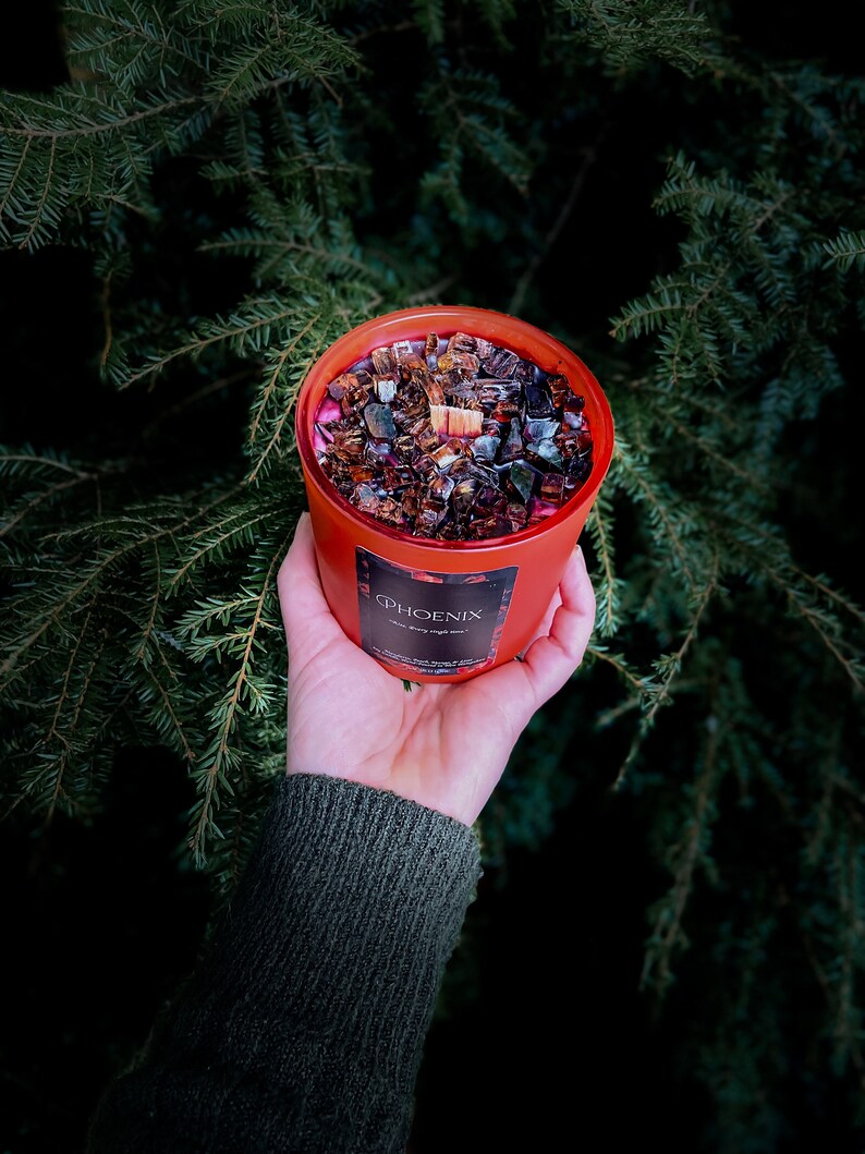 May include: A red candle jar with a black and brown wax blend and a wooden wick. The jar has a label that says "PHOENIX" with the words "with every single soul" underneath.