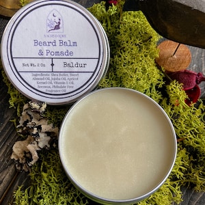 May include: A small tin of beard balm and pomade labeled "Sacred Lore Beard Balm & Pomade" with the name "Baldur" printed below. The tin is 2 ounces and the ingredients are listed on the label. The tin is sitting on a bed of green moss.