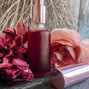 K&ouml;nnte beinhalten: Eine klare Glasflasche mit einer rosafarbenen Fl&uuml;ssigkeit und einem ros&eacute;goldenen Spr&uuml;hkopf. Die Flasche steht auf einer Holzoberfl&auml;che mit rosa und burgunderfarbenen Blumen.