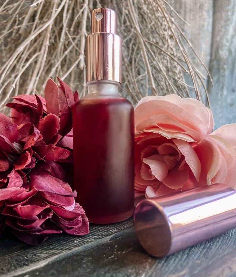 K&ouml;nnte beinhalten: Eine klare Glasflasche mit einer rosafarbenen Fl&uuml;ssigkeit und einem ros&eacute;goldenen Spr&uuml;hkopf. Die Flasche steht auf einer Holzoberfl&auml;che mit rosa und burgunderfarbenen Blumen.
