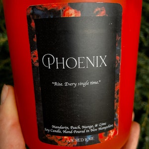 May include: Red glass candle jar with a black label that reads "PHOENIX" and "Rise. Every single time." The label also lists the scent as "Mandarin, Peach, Mango, & Lime" and states that the soy candle is hand-poured in New Hampshire. The label also includes the brand name "SACRED LORE".