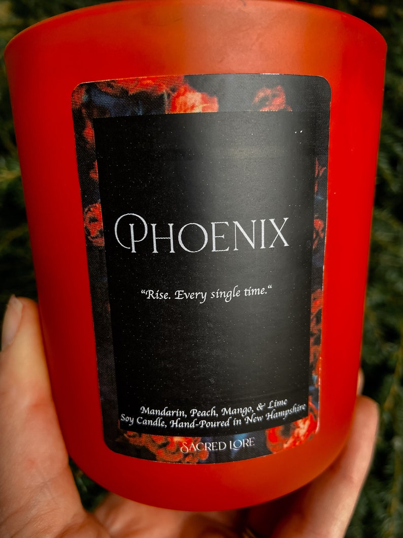 May include: Red glass candle jar with a black label that reads "PHOENIX" and "Rise. Every single time." The label also lists the scent as "Mandarin, Peach, Mango, & Lime" and states that the soy candle is hand-poured in New Hampshire. The label also includes the brand name "SACRED LORE".