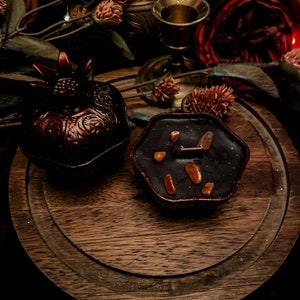 Puede incluir: Una bandeja de madera marr&oacute;n oscuro con una caja decorativa en forma de granada y una vela negra con trozos de cera naranja. La vela tiene forma de hex&aacute;gono.