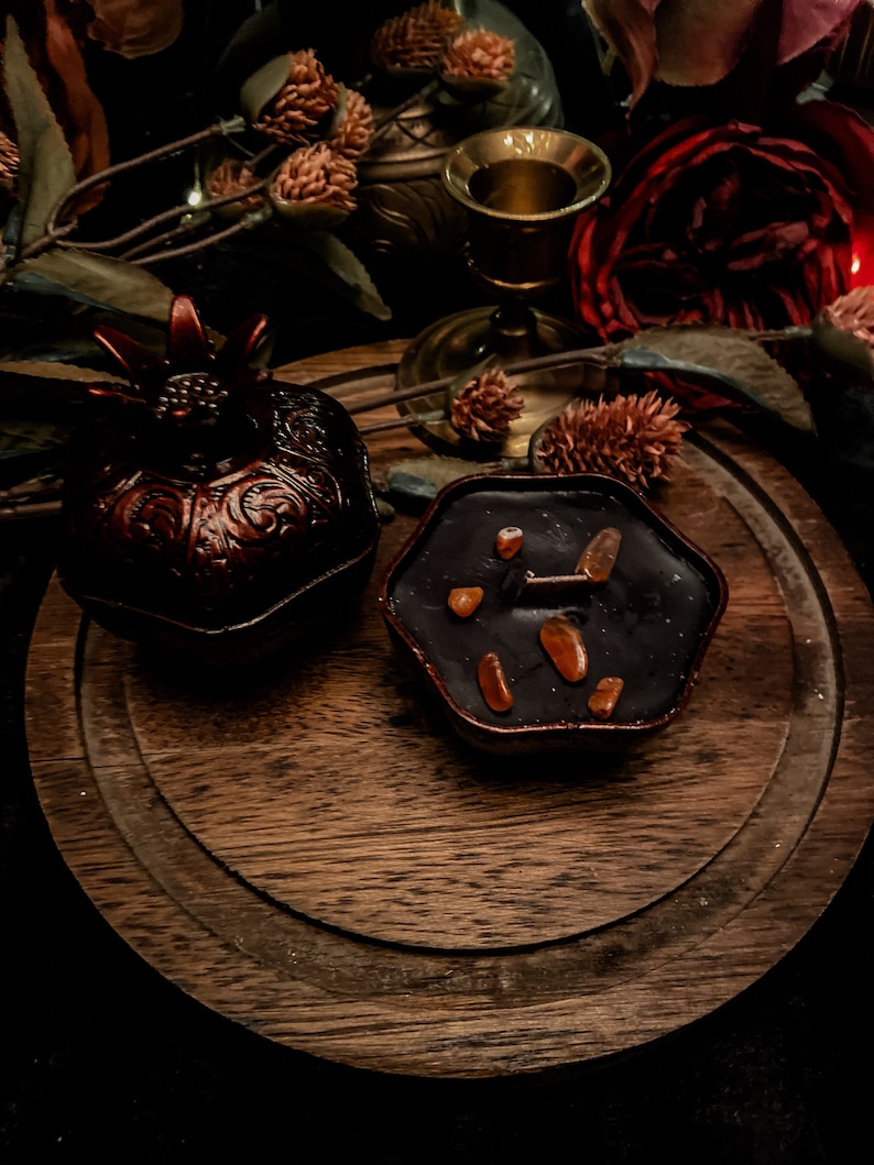 Puede incluir: Una bandeja de madera marr&oacute;n oscuro con una caja decorativa en forma de granada y una vela negra con trozos de cera naranja. La vela tiene forma de hex&aacute;gono.