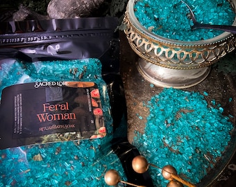 May include: A bag of teal blue bath salts with the text "Sacred Lore Feral Woman Ritual Bath Soak" on the front. The bath salts are in a clear glass bowl with a gold metal rim.