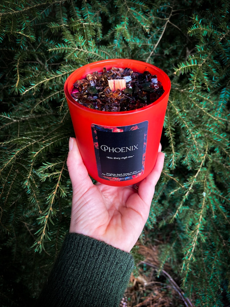 May include: Red glass candle jar with a black label that says "PHOENIX" and "Rise. Every single time." The jar is filled with brown wax and has a wooden wick.