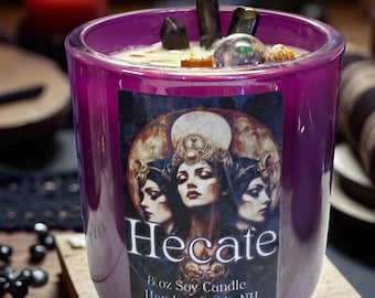 Puede incluir: Un recipiente de vela de vidrio morado lleno de cera, rematado con cristales oscuros y una pequeña esfera. La etiqueta presenta una ilustración de tres rostros y el texto Hecate, 8 oz Soy Candle, Handcrafted in NH.