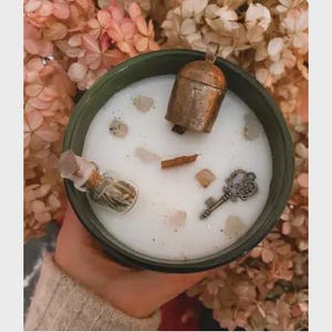 May include: A round dark green candle container filled with white wax. Embedded in the wax are a small metallic bell, a miniature glass bottle containing dried herbs, a metal key, and several small light-colored stone fragments.