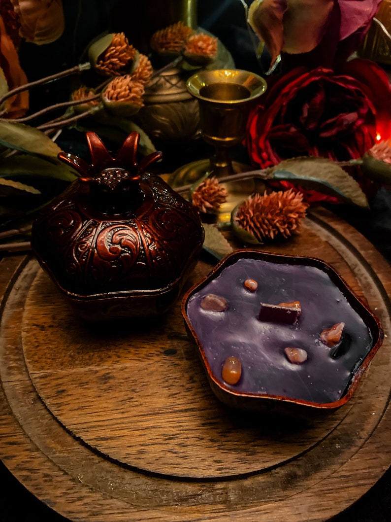 Puede incluir: Una vela morada con motas marrones en un recipiente con forma de granada. La vela est&aacute; sobre una superficie de madera con flores secas y un candelero de metal dorado.